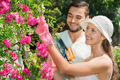 A woman and a man are working together in a lush garden, pruning a vibrant pink flowering shrub with a pair of garden clippers. The woman, wearing a white sun hat and pink polka-dot gardening gloves, is reaching up to trim the plant, while the man, dressed in casual clothing with a beard, stands beside her holding a small garden fork. The garden features well-maintained flower beds with dense greenery, including a hedge lining the background and a variety of healthy shrubs and trees that provide natural shade. The scene is outdoors on a bright, sunny day with clear weather, capturing natural light illuminating the green foliage and colorful blossoms. This image exemplifies professional outdoor gardening and landscaping activities suitable for service providers such as Lawn Mowing Sydenham, emphasizing the importance of garden upkeep, pruning, and plant care in a landscaped residential environment in Sydenham or nearby area, aligning well with gardening service offerings.
