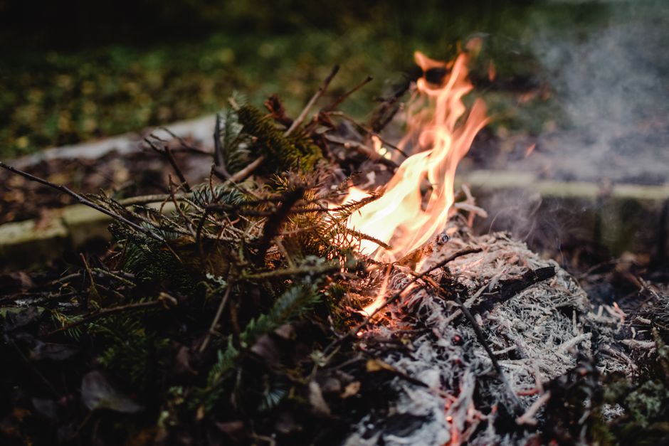 A close-up view of a garden bonfire within a large, round metal fire pit, with several stacked wooden logs ignited and producing bright orange flames and some wisps of smoke. The fire pit is situated