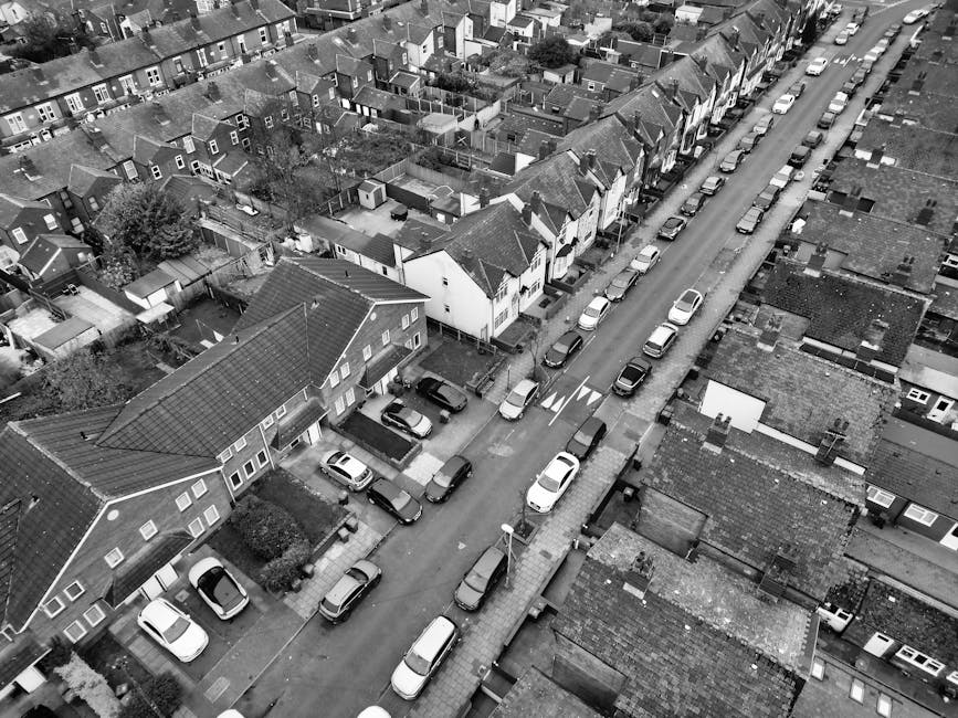 An aerial black-and-white view of a residential street featuring a row of semi-detached houses with pitched roofs and small front gardens, some with hedges or trees. A narrow, paved road runs through