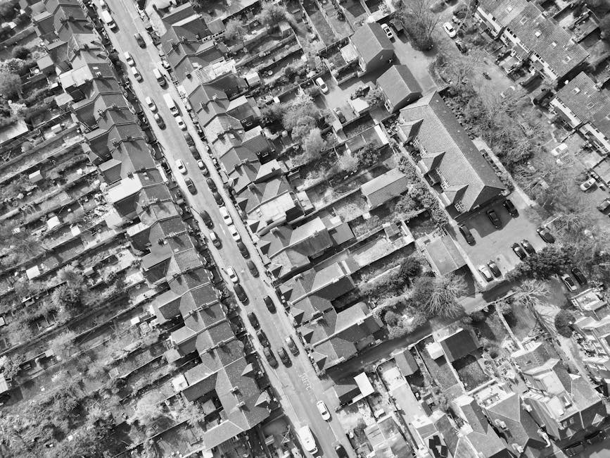 An aerial black-and-white view of a residential street featuring a row of semi-detached houses with pitched roofs and small front gardens, some with hedges or trees. A narrow, paved road runs through